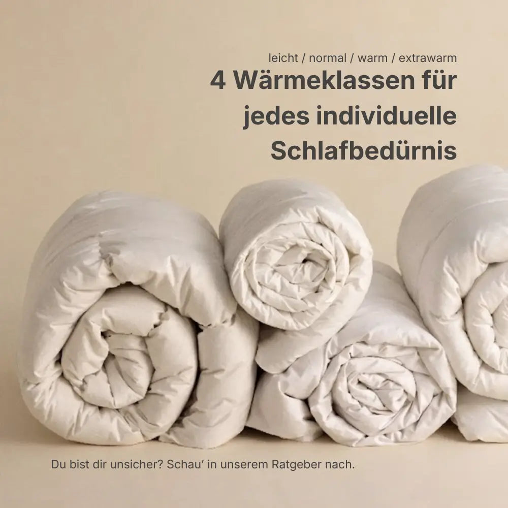 Mehrere zusammengerollte Daunendecken nebeneinander, Hinweis auf vier Wärmeklassen – leicht, normal, warm und extrawarm – für individuelles Schlafbedürfnis