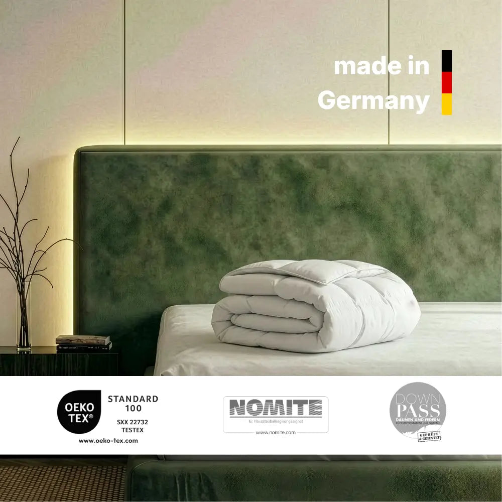 Zusammengelegte Superior Daunendecke auf einem Bett mit grünem Kopfteil, Hinweis ‚Made in Germany‘ sowie Zertifikate OEKO-TEX Standard 100, NOMITE und Downpass sichtbar