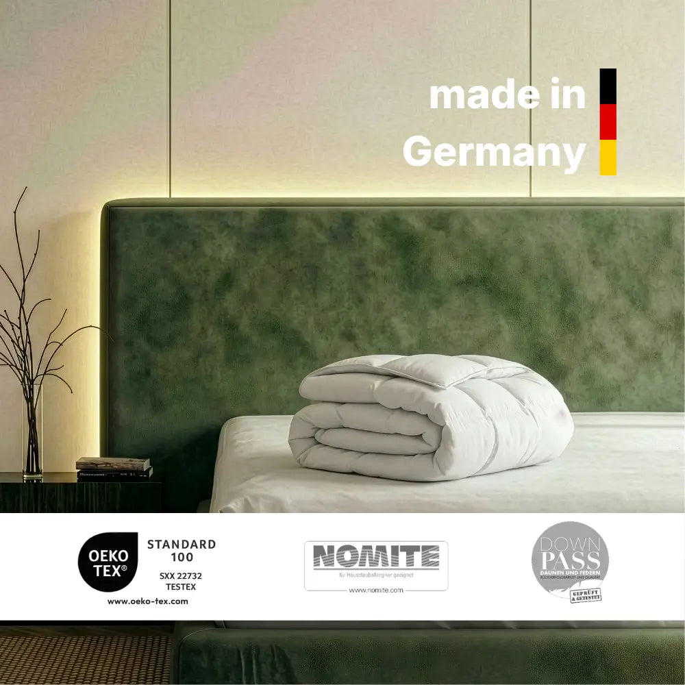 Zusammengelegte Superior Daunendecke auf einem Bett mit grünem Kopfteil, Hinweis ‚Made in Germany‘ sowie Zertifikate OEKO-TEX Standard 100, NOMITE und Downpass sichtbar